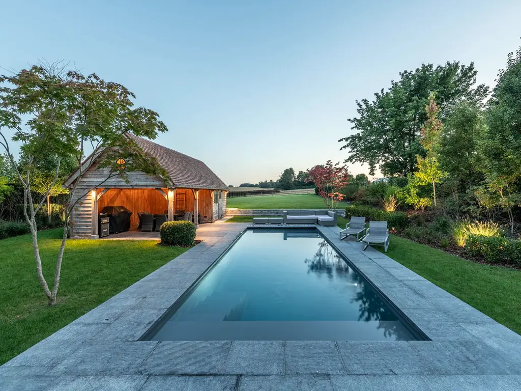 Lang zwembad en houten poolhouse met verlichte zithoek bij schemering — Klassieke tuin met zwembad, Roosdaal