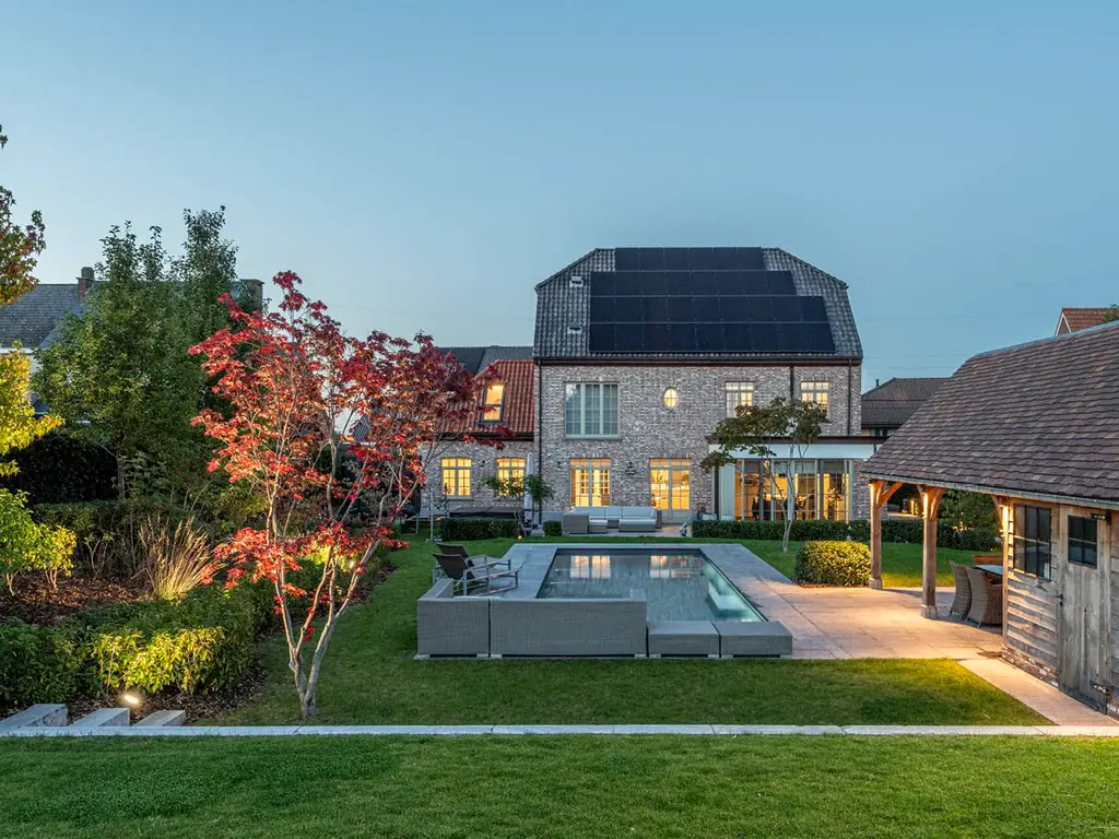 Zwembad met loungeset, houten overkapping en verlichte woning in de schemering — Klassieke tuin met zwembad, Roosdaal