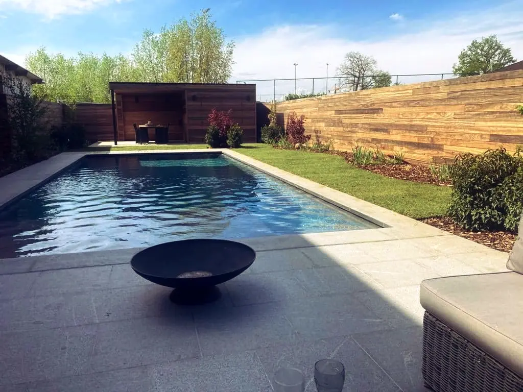 Lang zwembad met lichte rand, vuurschaal en loungezetel, houten poolhouse achteraan — Oase-tuin, Ternat