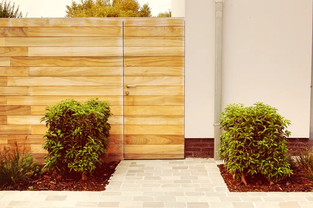 Houten poort in een houten wand, pad van lichte kasseien en twee bolvormige heesters — Open en toch gezellig