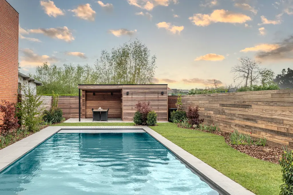 Langwerpig zwembad met houten poolhouse, houten schutting en gazon bij avondlicht