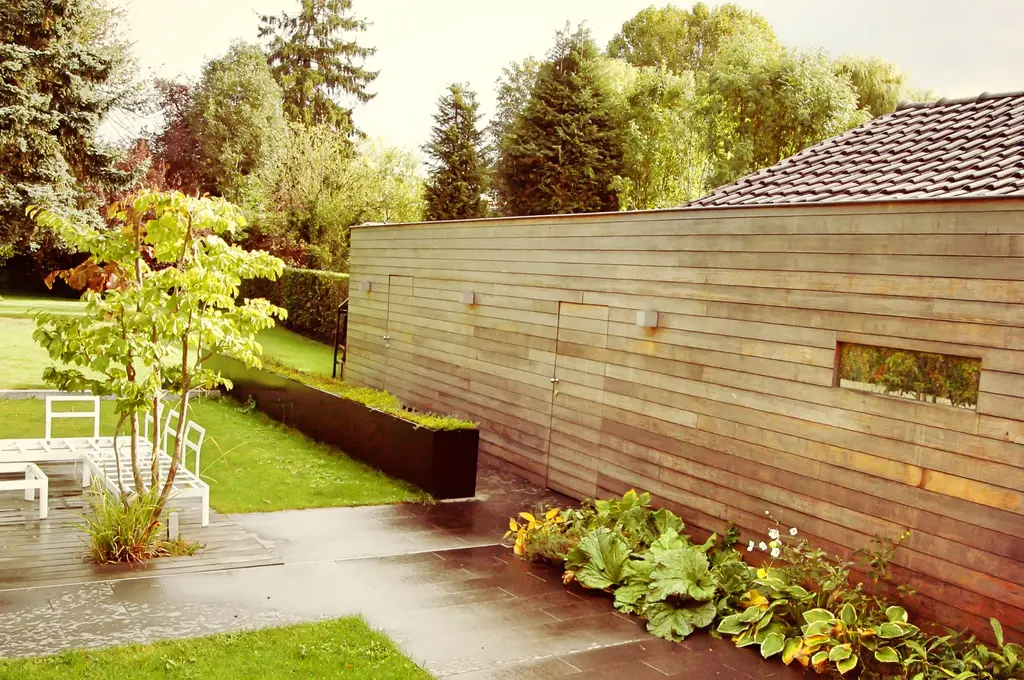Lange houten wand met smal raam, donkere tegels, gazon en witte tuinstoelen — Smeltkroes van stijlen, Dilbeek