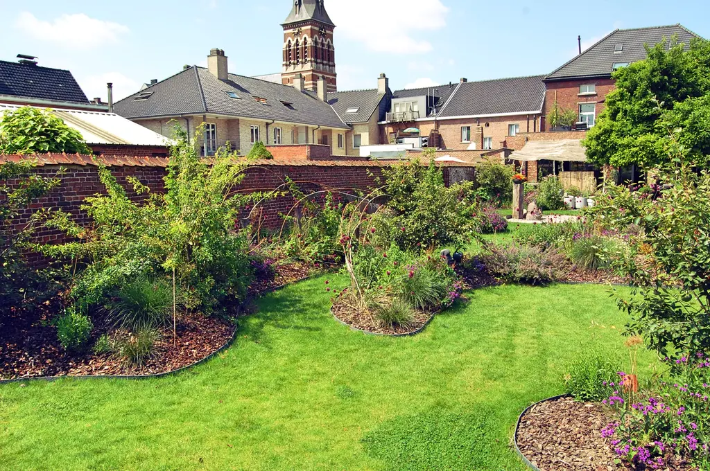 Gazon met golvende plantvakken langs een bakstenen muur, kerktoren in de verte — Speelse taluds, Galmaarden